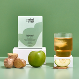 Green-themed product set with 'mini + me' packaging, apple, ginger, and a glass of juice on a green background.