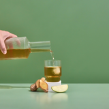 Hand pouring a clear bottle of juice into a glass with ginger root and apple slices on a green background