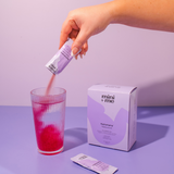 Hand pouring a pink powder from a 'mini + me' packet into a glass on a purple surface.