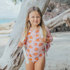 Young girl in a strawberry-patterned swimsuit standing by a tree on a beach.