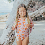 Young girl in a strawberry-patterned swimsuit standing by a tree on a beach.