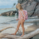 Person in a pink swimsuit with orange patterns standing on a log at the beach.