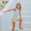 Young girl in a striped swimsuit and shorts sitting on a white surface.