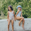 Two young girls in swimsuits sitting on a concrete ledge with a green bush background