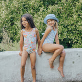 Two young girls in swimsuits sitting on a concrete ledge with a green bush background