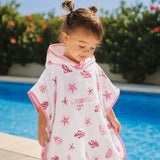 A toddler wearing a white hooded towel poncho with pink trim and red starfish and shell prints, standing by a swimming pool outdoors. The brand name CRANVOLE is printed on the front.