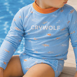 A close-up of a baby wearing a light blue long-sleeve swim shirt with a fish pattern and the brand name 'CRYWOLF' printed in white across the chest. The baby is sitting by a swimming pool.