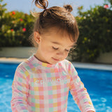 A young child wearing a pastel checkered long-sleeve swim shirt with the brand name 'CRYWOLF' printed across the chest, standing by a swimming pool with green foliage in the background.