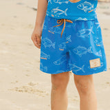 A child wearing bright blue swim trunks and a matching top printed with white fish, standing on a sandy beach. The shorts have an orange drawstring and a small tan brand patch on the lower right leg.