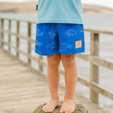 A child standing barefoot on a wooden dock wearing bright blue swim trunks with a white fish pattern and a light blue shirt. The background shows a blurred body of water and more dock railings.