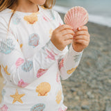 A child wearing a white long-sleeve rash guard with a colorful seashell and starfish print, holding a pink seashell on a beach.