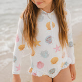 A young girl wearing a white long-sleeve swim shirt with a half-zip front and an all-over seashell and starfish print, standing on a sandy beach with the ocean in the background.