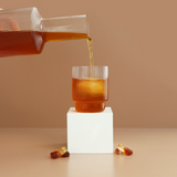 Glass pitcher pouring amber liquid into a glass on a beige background