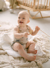 Baby sitting on a textured blanket holding a wooden toy, with a crib in the background.