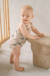 Baby in a beige romper standing next to a beige crib in a softly lit room.