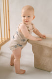 Baby in a beige romper standing next to a beige crib in a softly lit room.