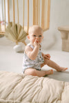 Baby sitting on a beige blanket in a room with wooden furniture and a white wall.