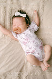 Newborn baby wearing a floral onesie and headband, lying on a soft surface.