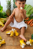 Child sitting on a wooden floor surrounded by vegetables and fruits, eating a piece of fruit.