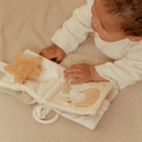 Baby playing with a soft book featuring star and moon illustrations on a beige surface.
