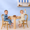 Two children sitting at a table with wooden chairs, playing with toys against a blue wall.