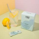 Glass of lemonade with a straw, lemon slices, and 'mini + me' product packaging on a pastel background.