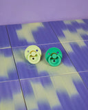 Two pacifiers, one green and one yellow, on a blue and purple checkered surface.
