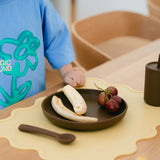 Child's hand reaching for a banana on a plate with grapes and a spoon on a table.