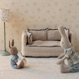 A styled dollhouse or miniature room scene with a small beige sofa, a floral-patterned floor lamp, and two plush bunny toys on a cream rug against floral wallpaper.