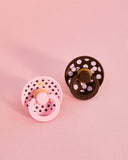 Two baby pacifiers, one pink and one brown with polka dot design, on a pink background.
