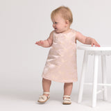 Baby in a light pink dress standing next to a white stool on a plain background