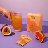 Hand holding a glass of orange juice with a 'mini + me' box and packets on a purple background