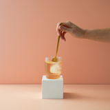 Hand stirring a glass of iced tea with a spoon on a peach-colored background