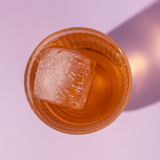 Glass of orange liquid with a large ice cube on a light purple background