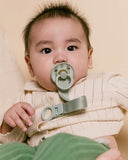 Baby wearing a beige outfit with a green pacifier and strap against a beige background
