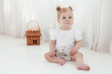 Baby sitting on a white surface wearing a white onesie with a floral diaper cover, next to a small woven basket.