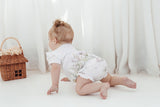 Baby in a white outfit with floral patterns sitting on a white floor with a wicker basket.