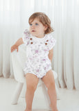 Baby wearing a floral romper with brown buttons against a white curtain background