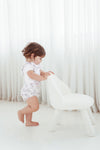 Child in a white romper standing next to a white chair against a white curtain background