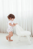 Child in a white romper standing next to a white chair against a white curtain background