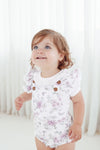 Child wearing a floral onesie with brown buttons against a white curtain background