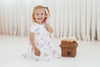Child in a white floral dress sitting next to a small wicker basket on a white background