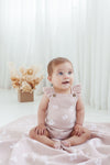 Baby in a pink floral romper sitting on a white blanket with a neutral-colored curtain background.