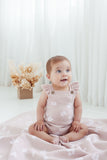 Baby in a pink floral romper sitting on a white blanket with a neutral-colored curtain background.