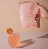 Hand holding a pink package labeled 'franjos kitchen' with a glass of pink drink and orange slice on a beige background.
