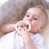 A baby lying on their back, holding a cream-colored textured teether toy in their mouth and hands, set against a soft, patterned beige bassinet.