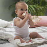 A baby sitting on a bed holding a Sophie De Luxe giraffe teething toy, wearing a white sleeveless bodysuit.