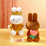 Two plush toys, one white with a crown and yellow outfit, the other brown with a pink flower, on a beige background.