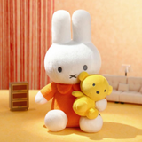 Plush toy rabbit wearing an orange outfit holding a yellow teddy bear in a room with a striped curtain and sofa.