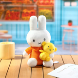 Plush toy rabbit in an orange shirt holding a yellow teddy bear on a wooden floor.